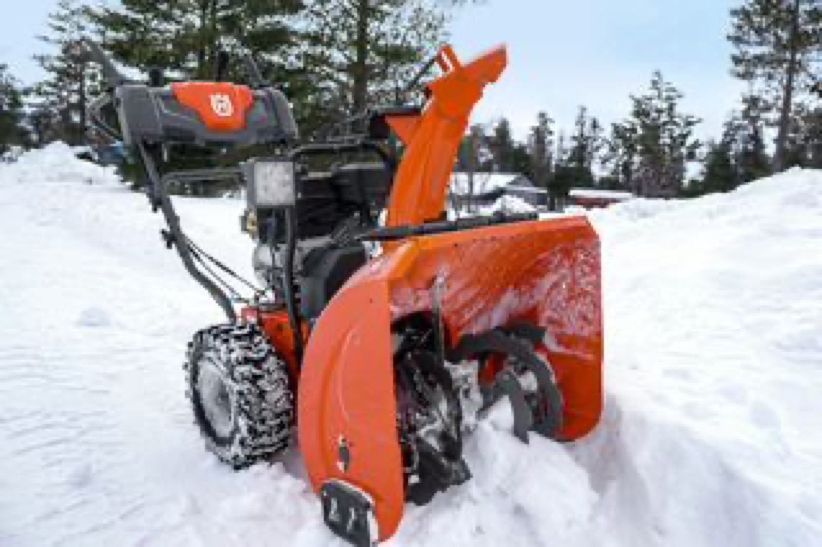 Nærbilde av en oransje totrinns Husqvarna snøfreser i snøen med furutrær og lys himmel bak